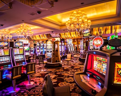 A vibrant casino interior filled with rows of colorful slot machines, each illuminated with bright lights and displays. The ceiling is adorned with elegant chandeliers and golden drapes, creating a luxurious atmosphere. The patterned carpet adds to the elaborate design of the space.